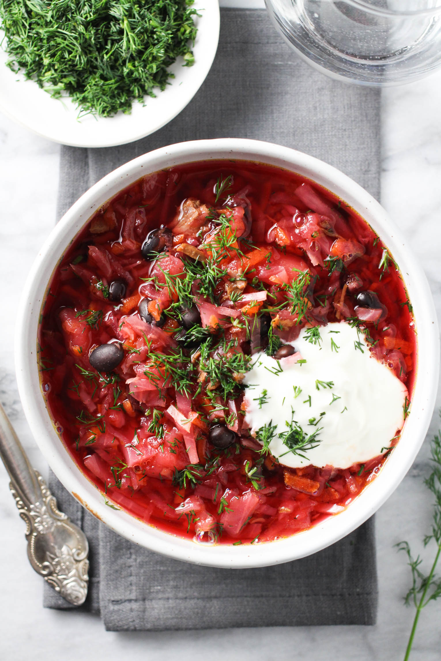 Overhead shot of borscht in a bowl standing on a grey napkin. The soup is garnished with sour cream and chopped dill.