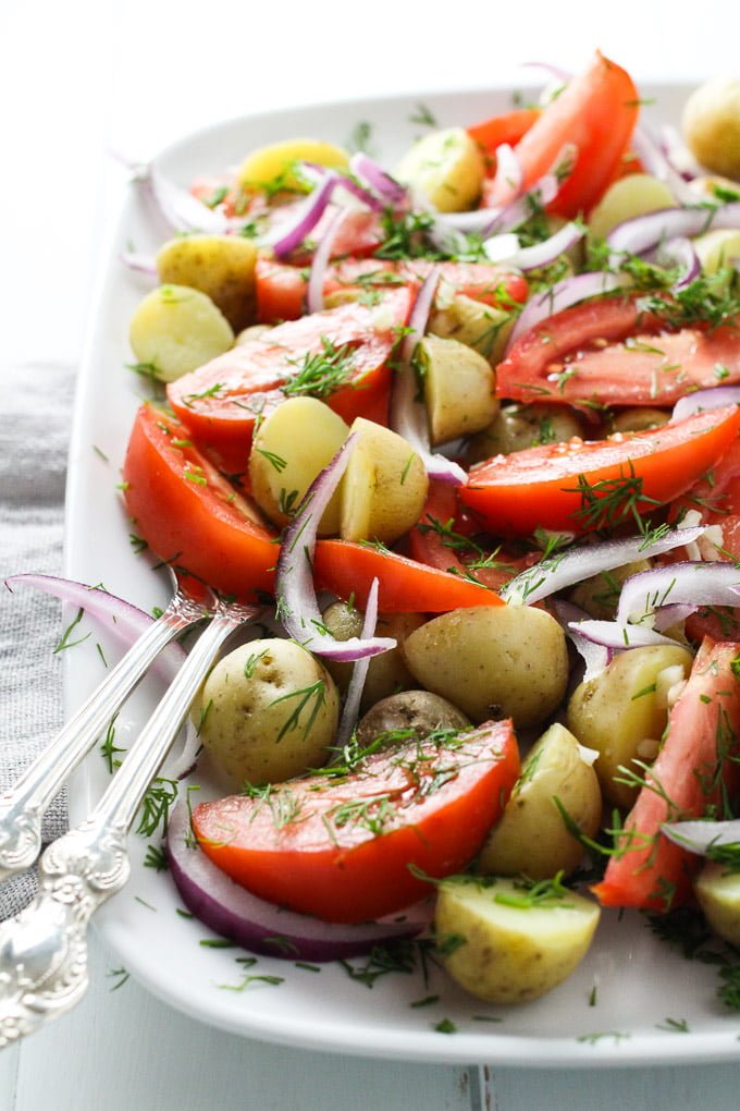 New potato salad with tomatoes and dill on a white platter. Side view.