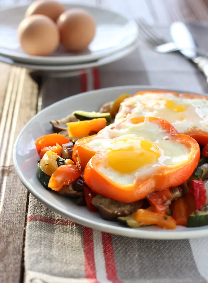 Two fried eggs over roasted vegetables on a plate. Three whole eggs in the backgound on white plates.