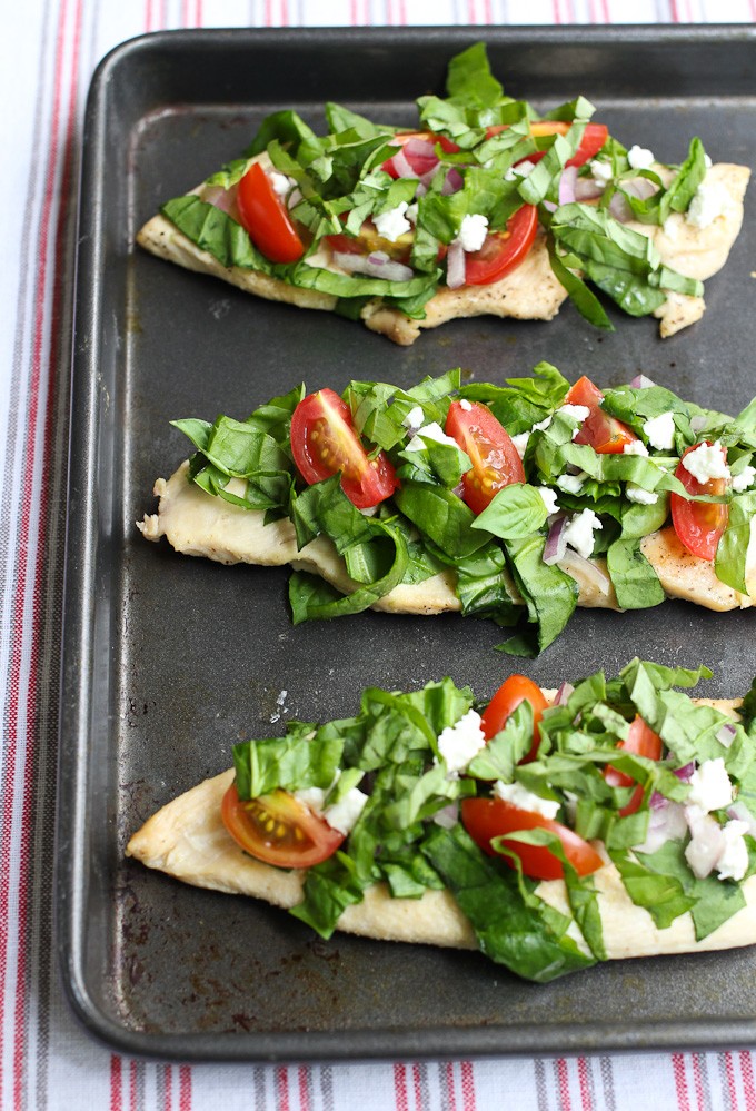 Chicken topped with spinach, cherry tomatoes, goat cheese, and basil on a baking sheet.