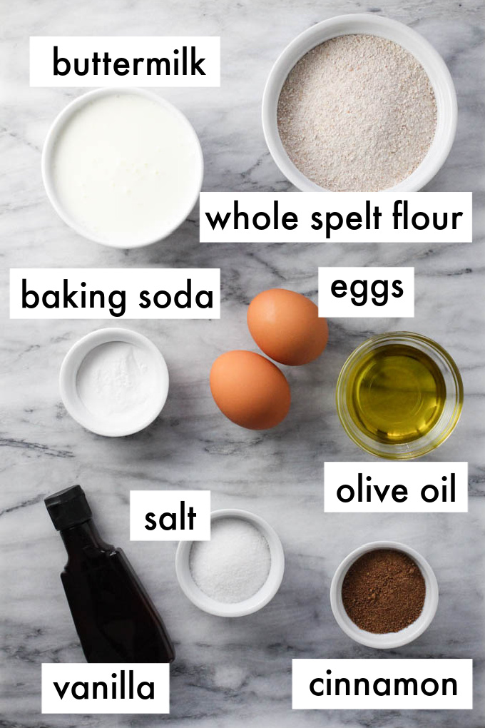 Spelt pancakes ingredients displayed on marble background. The ingredients are labeled as follows: buttermilk, whole spelt flour, baking soda, eggs, salt, olive oil, vanilla, cinnamon.