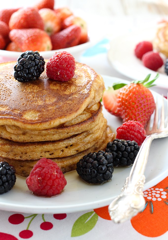 Spelt pancakes on a white plate, garnished with berries and maple syrup. Silver fork to the left. Strawberries in the background.