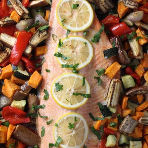 Baked rainbow trout fillet with roasted vegetables around it. The fish is garnished with lemon slices.