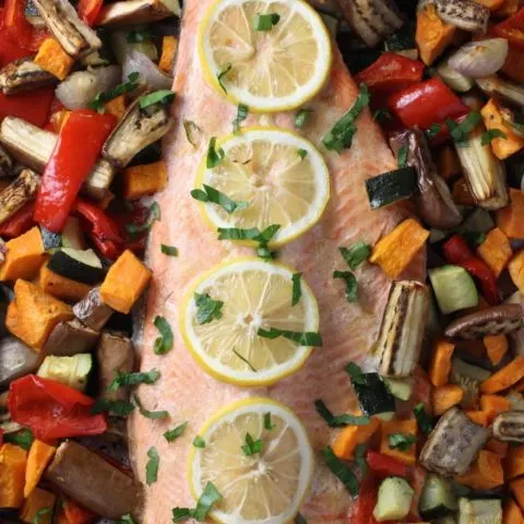 Baked rainbow trout fillet with roasted vegetables around it. The fish is garnished with lemon slices.