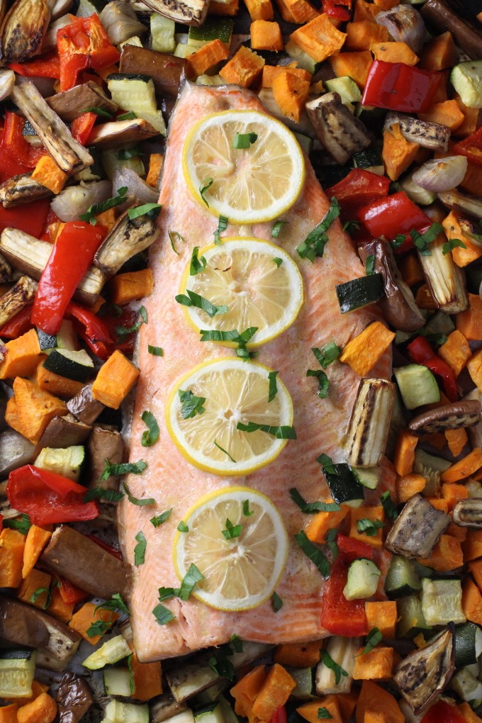 Baked rainbow trout fillet with roasted vegetables around it. The fish is garnished with lemon slices.