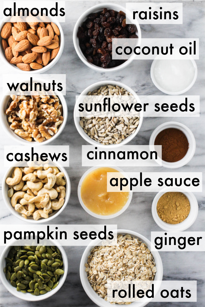 Granola ingredients displayed on marble background. The ingredients are labeled as follows: almonds, raisins, coconut oil, walnuts, sunflower seeds, cashews, cinnamon, apple sauce, pumpkin seeds, ginger, rolled oats.