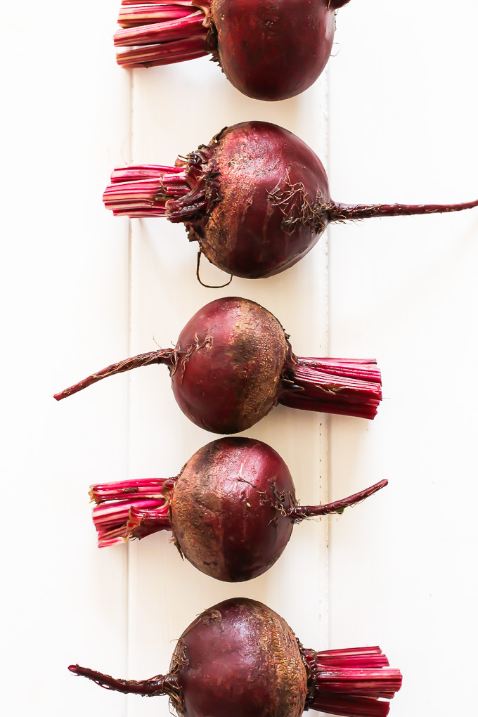 Six red beets with tops chopped off laying in a row on a white background. 