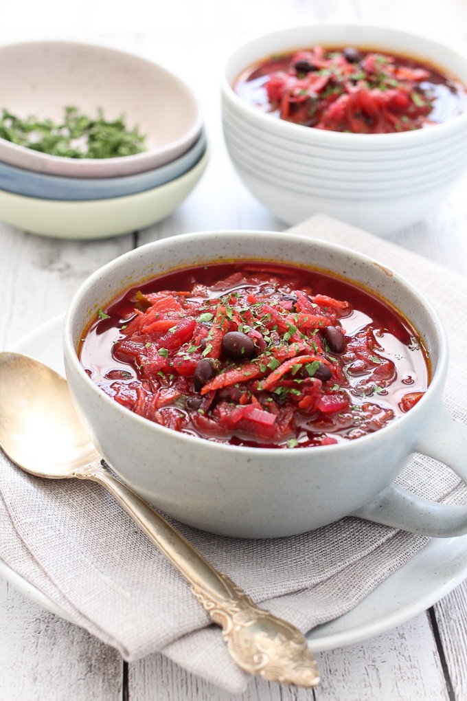 Beetroot soup with beans in a bowl with a spoon to the left. Another bowl of soup and chopped herbs in the background.