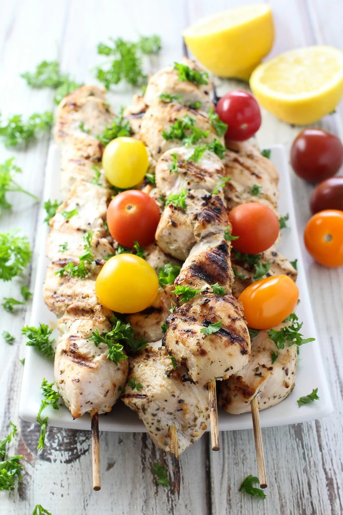 Yogurt marinated chicken kebabs on skewers, garnished with cherry tomatoes and chopped parsley. Lemon halves in the background.