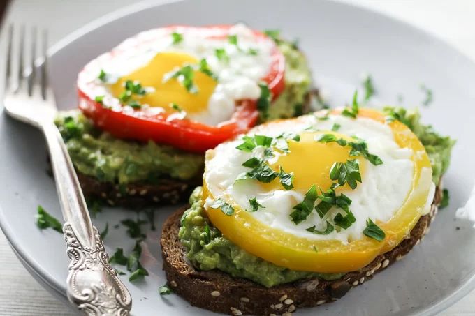 Close-up side view of two egg avocado toasts on a white plate with a silver fork on the left.