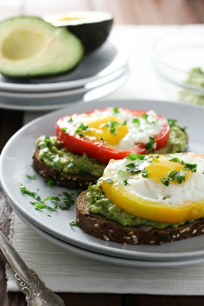 Close-up side view of two Egg Avocado Toasts on a plate with a sliced avocado in the background.