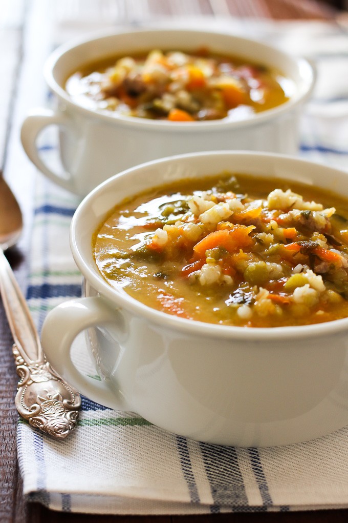 Close-up side view of the barley vegetable soup in a white bowl with handles standing on a kitchen towel. Second bowl is in the background. A silver spoon on the left.