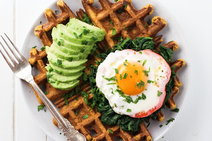 Top view of a sweet potato waffle topped with a fried egg, spinach and avocado. There is also a silver fork on the left.