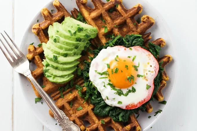 Top view of a sweet potato waffle topped with a fried egg, spinach and avocado. There is also a silver fork on the left.