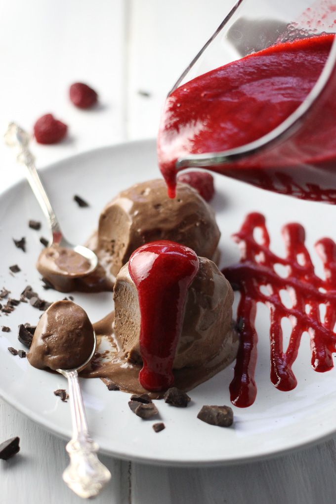 Raspberry sauce being poured over chocolate ice cream. Two silver spoons to the left of the ice cream.