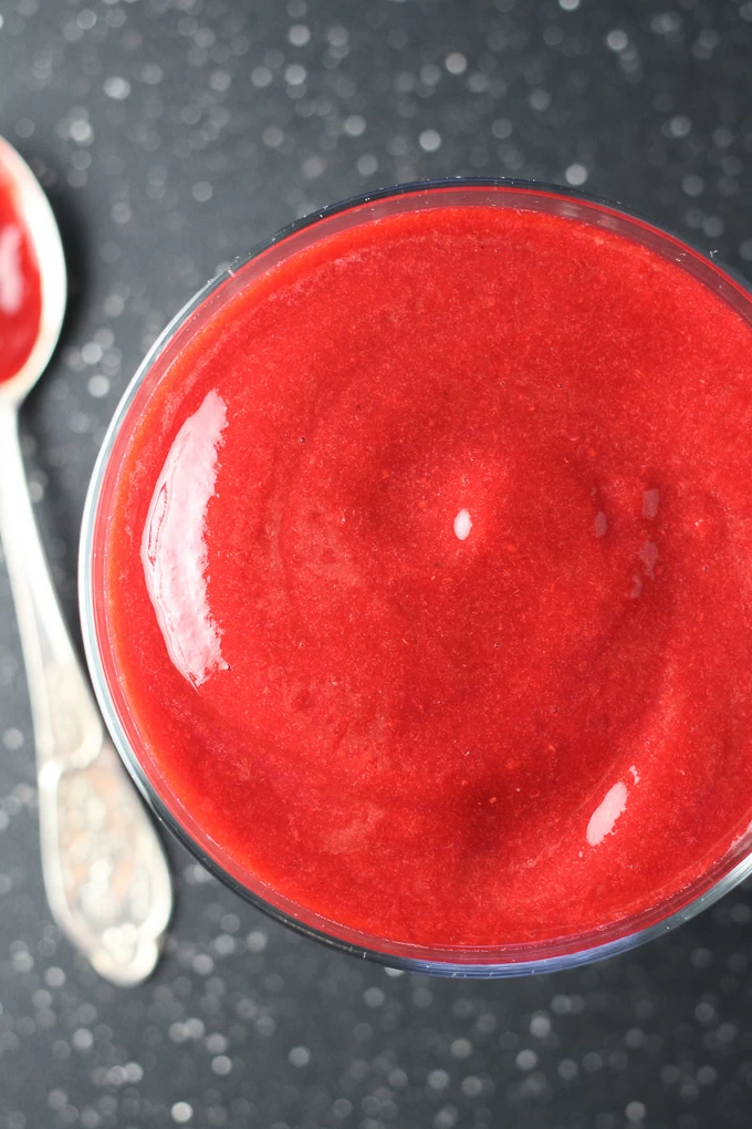 Top view of Raspberry Sauce in a glass standing on a dark background with a silver spoon on the left.