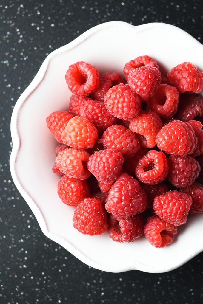Overhead shot of fresh red raspberries on a white plate standing on a dark background.