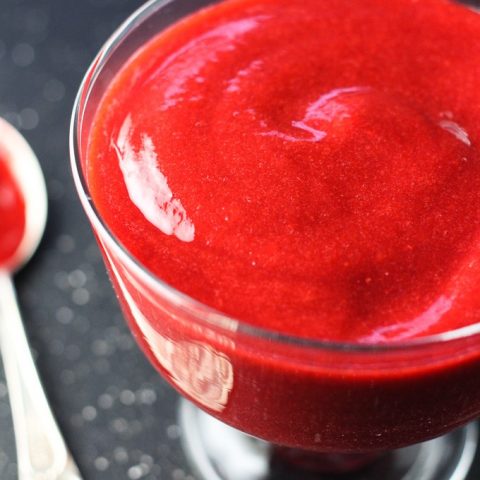 Close-up side view of the raspberry sauce in a glass on dark background with a silver spoon on the left.