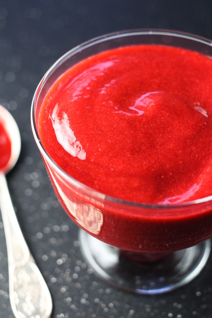 Close-up side view of the raspberry sauce in a glass on dark background with a silver spoon on the left.