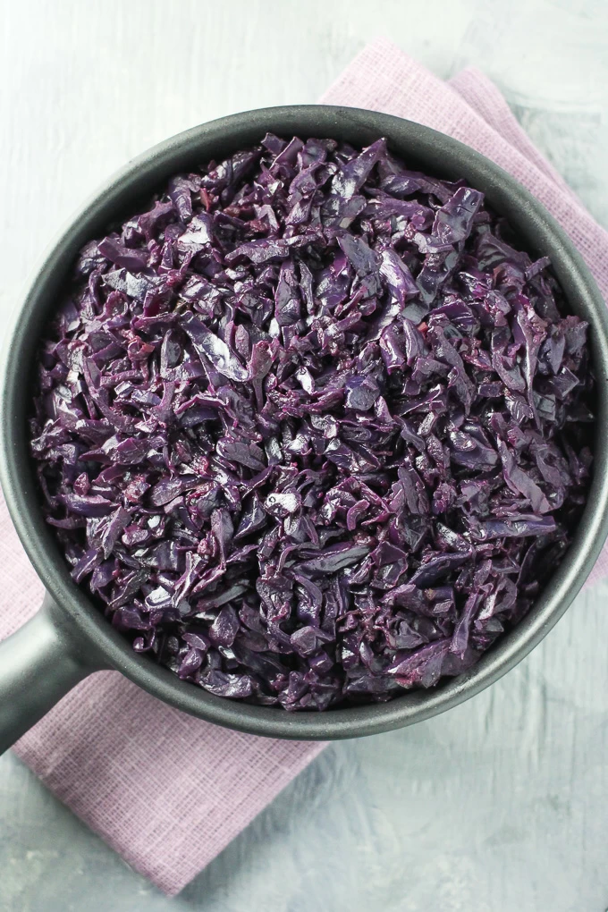 Top view of the braised German Red Cabbage in a black pan standing on a purple napkin on a grey background.