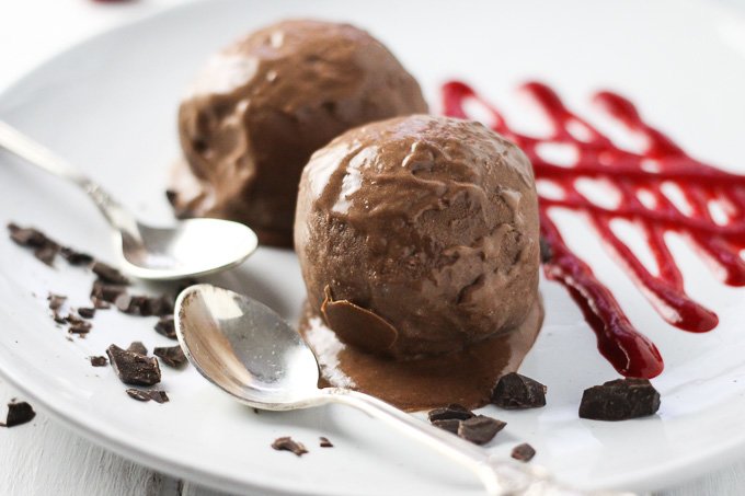 Close-up side view of the chocolate ice cream on a white plate with two silver spoons on the left and some raspberry sauce on the right.
