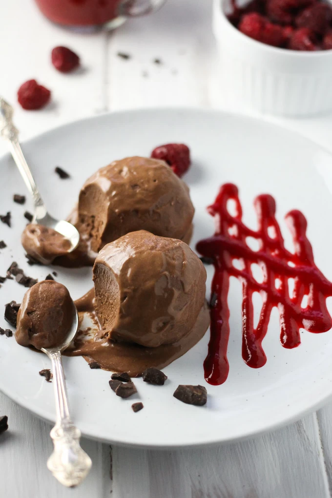 Close-up side view shot of the Vegan Chocolate Ice Cream on a white plate with two silver spoons on the left and some raspberry sauce on the right.