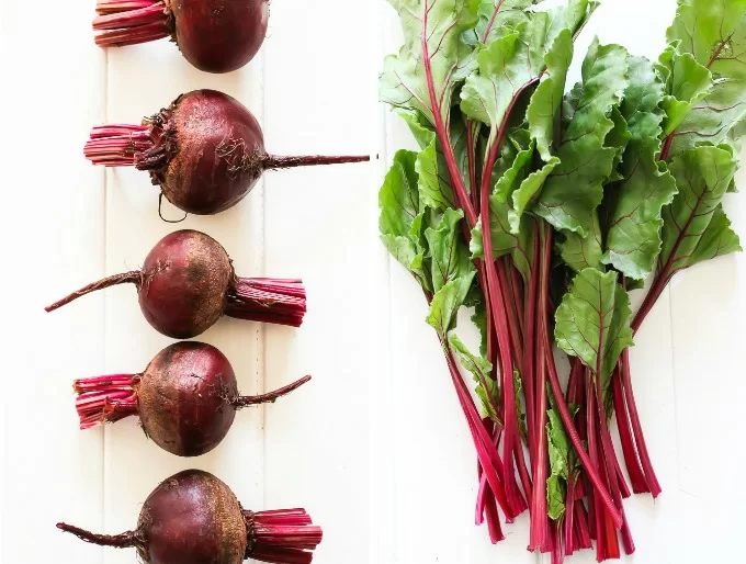 Two side-by-side top view images. On the left are beetroots with greens cut off, on the right is a bunch of beet greens.