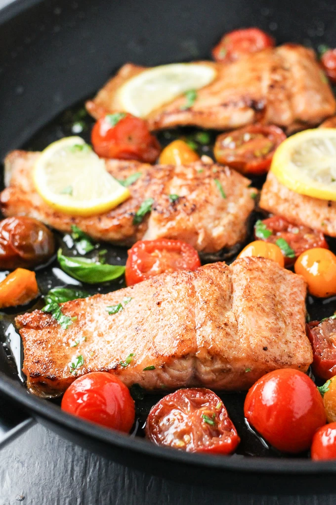 Close-up side view of the pan fried rainbow trout with cherry tomatoes and lemon slices in a black pan.