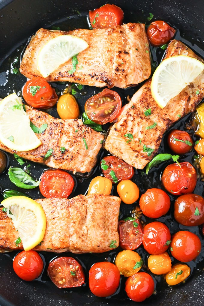 A top view of the pan fried rainbow trout with red cherry tomatoes and lemon slices in a black pan.