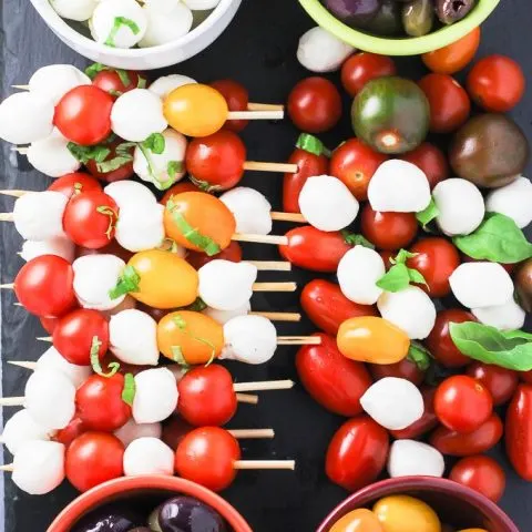 A top view of the caprese appetizer tray with the red and yellow cherry tomatoes, olives, small mozzarella cheese balls, and basil leaves arranged on a cheese board.
