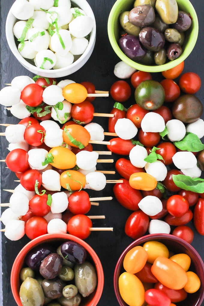 A top view of the caprese appetizer tray with the red and yellow cherry tomatoes, olives, small mozzarella cheese balls, and basil leaves arranged on a cheese board.