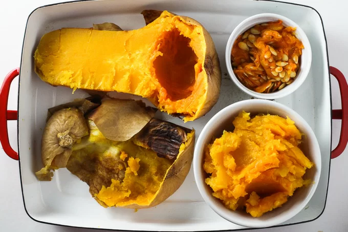How to Cook Butternut Squash: One half of a cooked butternut squash, a small dish with butternut squash seeds with pulp, skin of a cooked butternut squash, and a small dish with butternut squash puree, all placed inside a baking dish. 