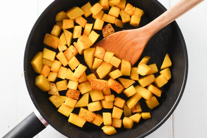 Top view of sauteed butternut squash cubes in a black pan with a wooden spatula in the middle.