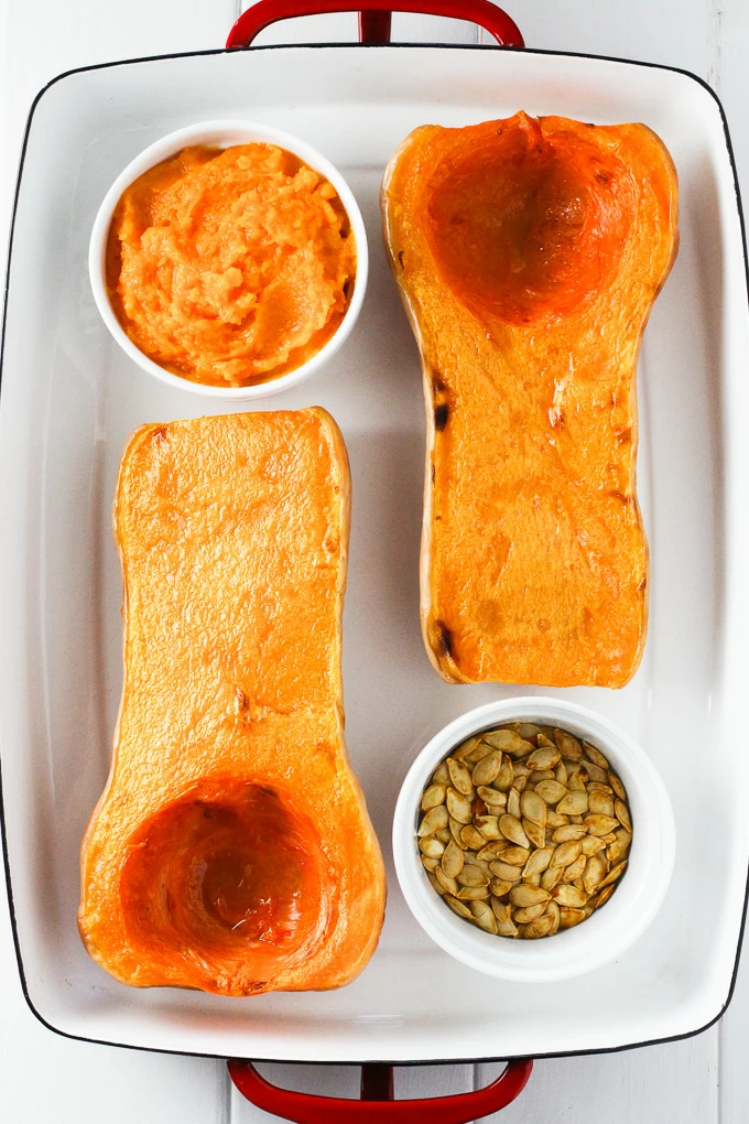 Two butternut squash halves, a small dish with butternut squash puree, and a small dish with butternut squash seeds, all placed into a white baking dish with red handles.