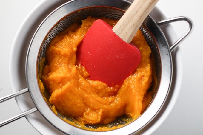 Top view of a butternut squash puree being pushed through a mesh colander placed over a white bowl. 