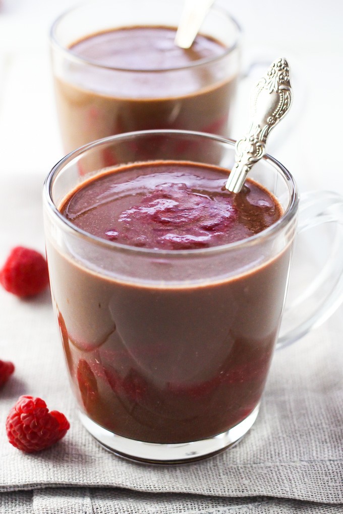 A close-up of the raspberry hot chocolate in a glass mug with another mug in the background and two raspberries on the left.