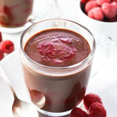 A close-up of the raspberry hot chocolate in a glass mug on a white plate with a spoon to the left and three raspberries on the right. Some raspberries and another mug in the background.