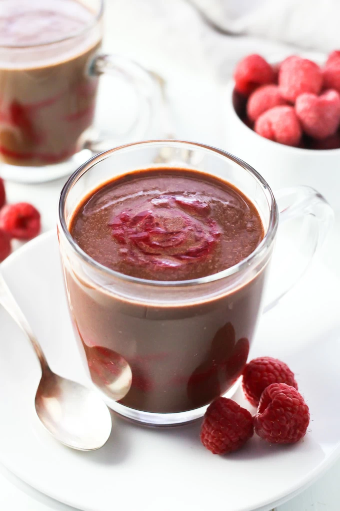 A close-up of the raspberry hot chocolate in a glass mug on a white plate with a spoon to the left and three raspberries on the right. Some raspberries and another mug in the background.