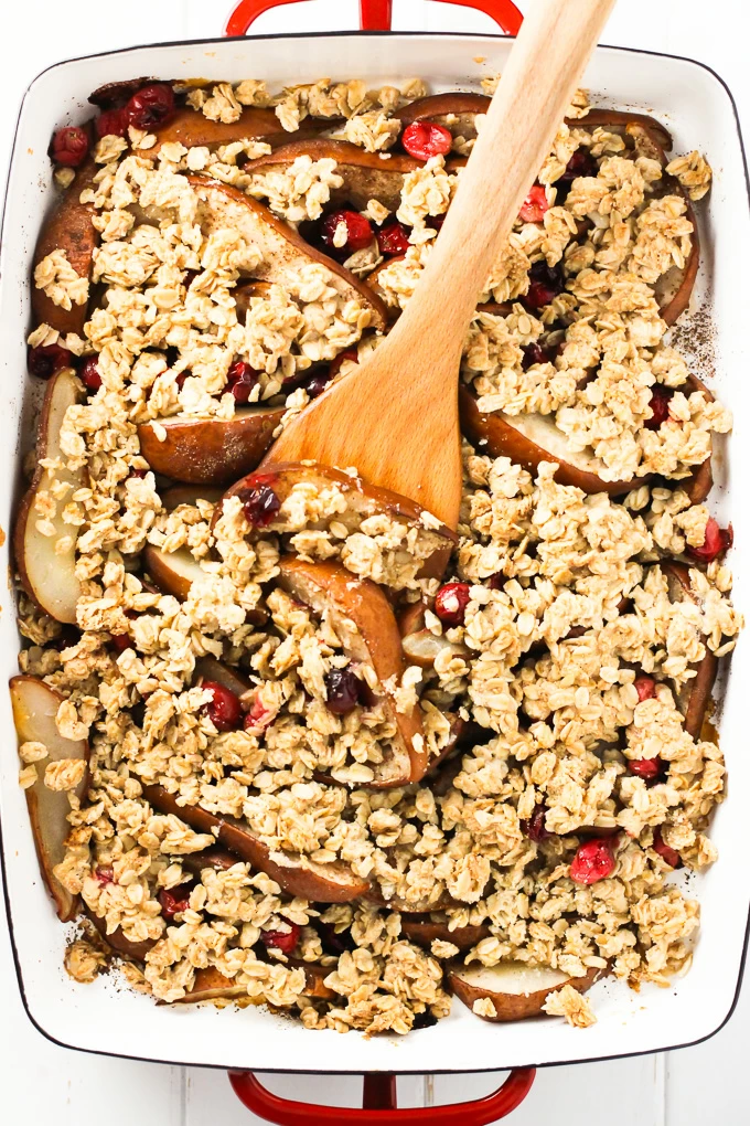 A close-up top view of a pear cranberry crisp with a wooden spatula in a white baking dish with red handles.