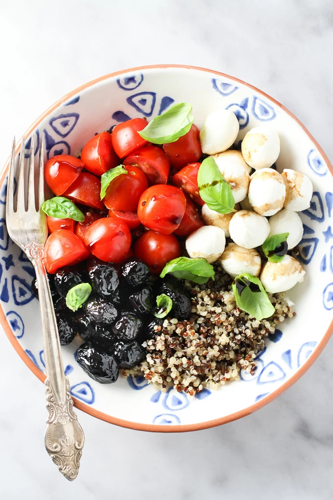 Caprese quinoa bowl with red cherry tomatoes, white bocconcini cheese, black olives, basil leaves and quinoa in a bowl with a silver fork on the left.