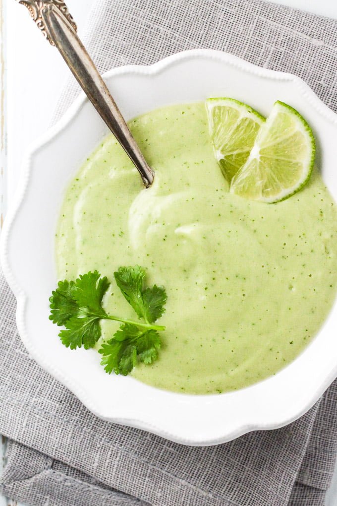 Cold avocado soup in a white bowl, garnished with cilantro and lime slices.