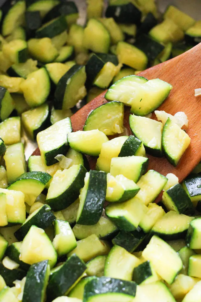 Close up of zucchini slices being tossed with a wooden spatula.