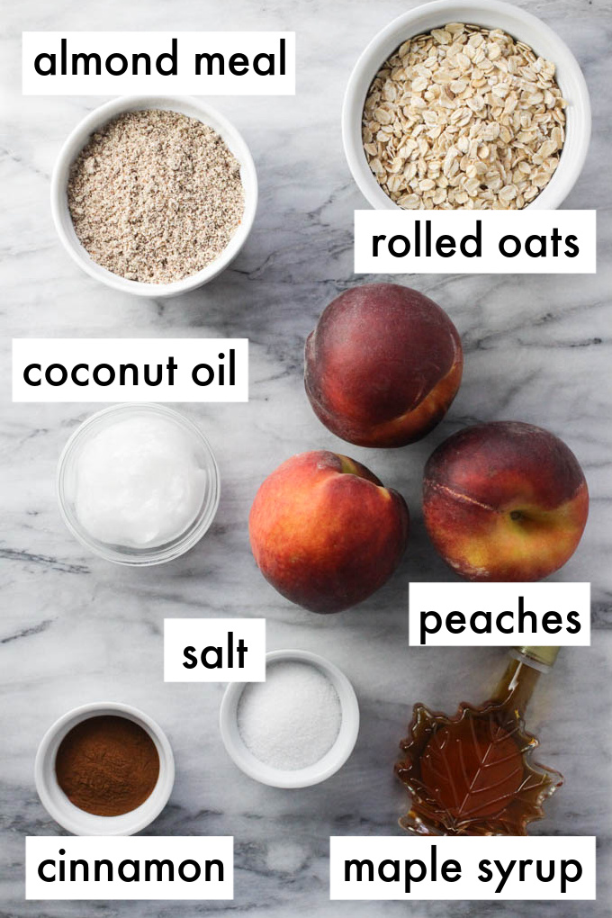 Ingredients for the peach crisp displayed on marble background. The ingredients are labeled as follows: almond meal, rolled oats, coconut oil, salt, peaches, cinnamon, maple syrup.