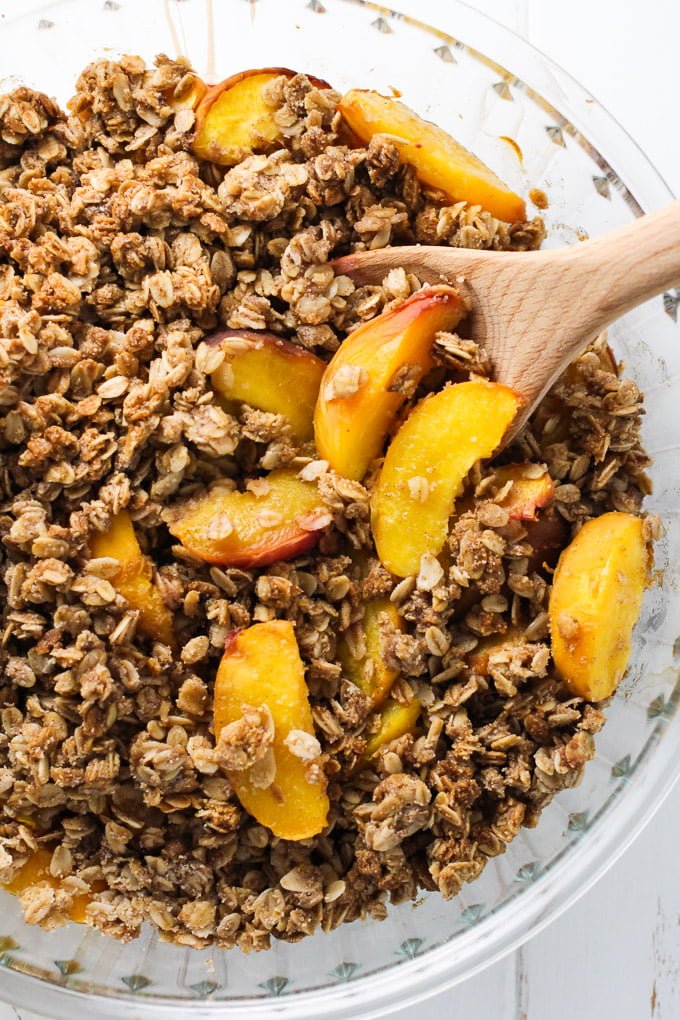 Healthy peach crisp in a round baking dish. The peach crisp is being scooped out with a wooden spoon.