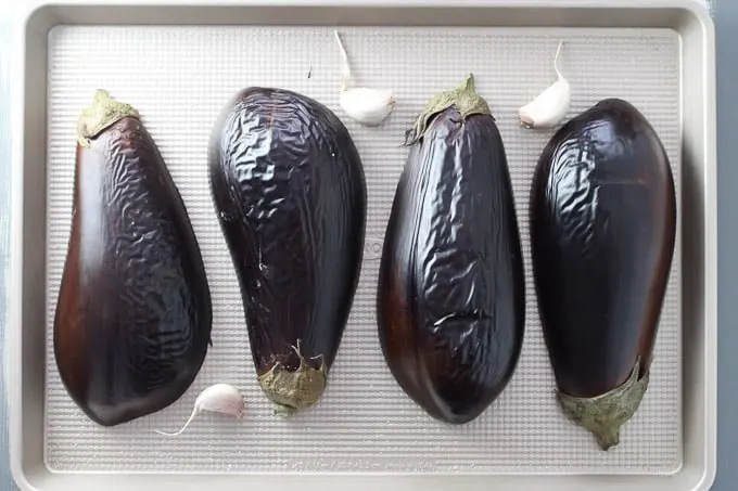 Slightly broiled eggplants on a baking sheet with three garlic cloves.