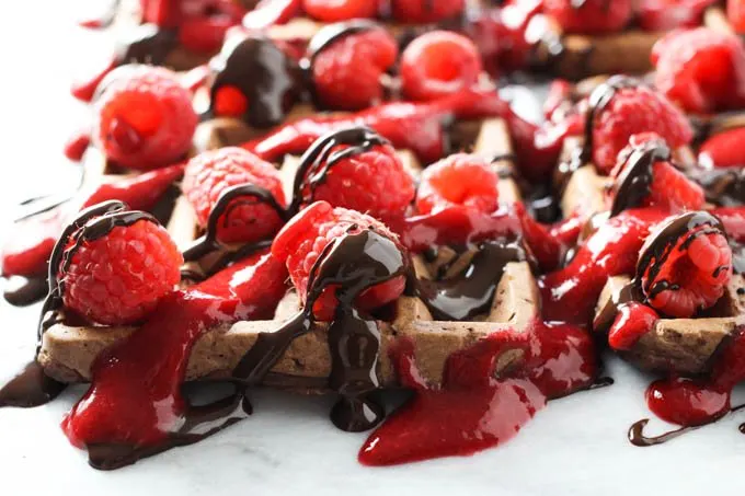 Close up of a chocolate waffle topped with fresh raspberries, chocolate sauce, and raspberry sauce. Side view.