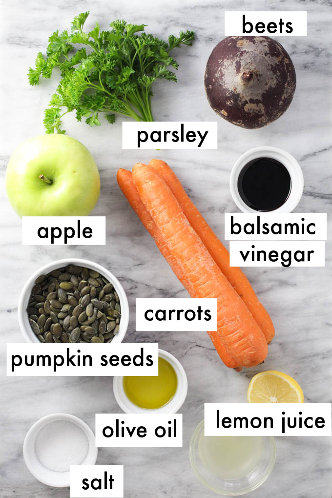 Ingredients for the raw beet salad on a marble background. The ingredients are labeled as follows: beets, parsley, apple, balsamic vinegar, carrots, pumkin seeds olive oil, lemon juice, salt.