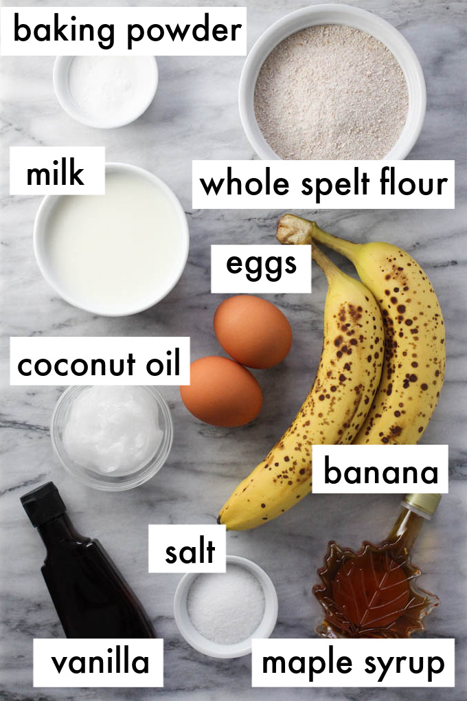 The ingredients for spelt banana bread dispayed on marble background. The ingredients are labeled as follows: baking powder, milk, whole spelt flour, eggs, coconut oil, salt, banana,vanilla, maple syrup.