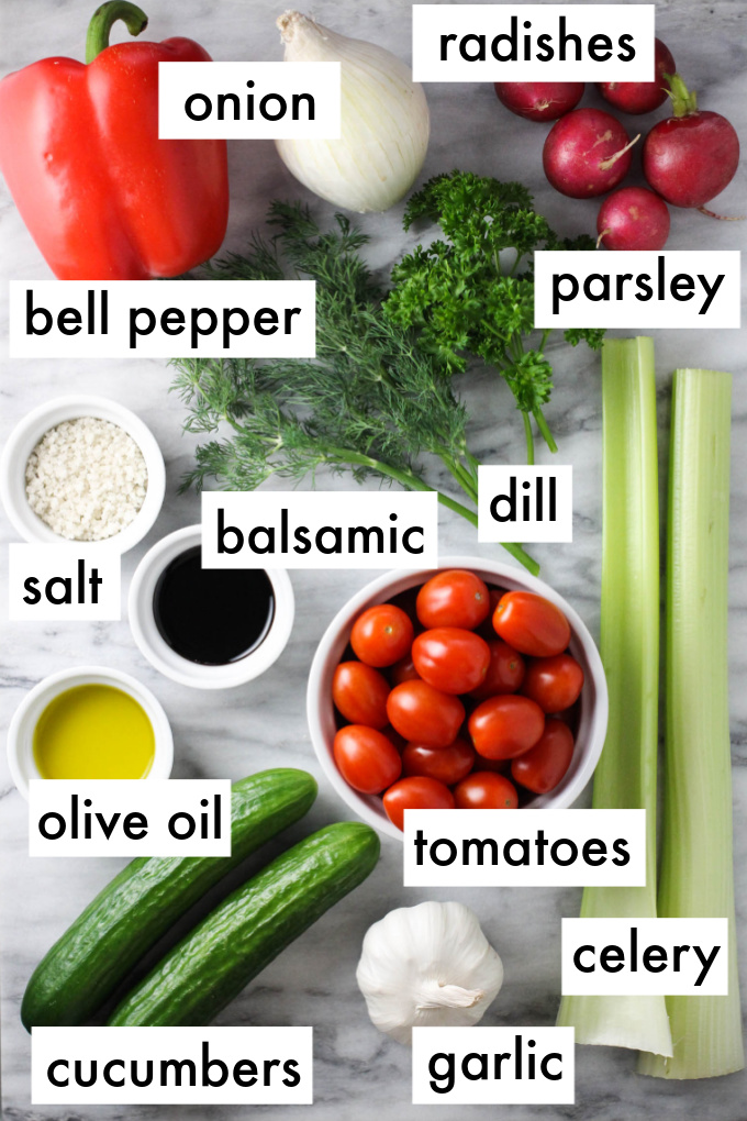 Ingredients for the salad displayed on marble background. The ingredients are labeled as follows: onion, radishes, bell pepper, parsley, salt, balsamic, dill, olive oil, tomatoes, celery, cucumbers, garlic.