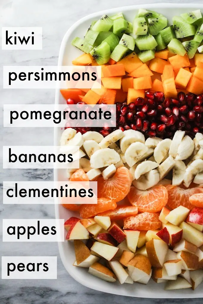 Ingredients for the winter fruit salad displayed in rows on a platter. Ingredients labels are as follows: kiwi, persimmons, pomegranate, bananas, clementines, apples, pears.